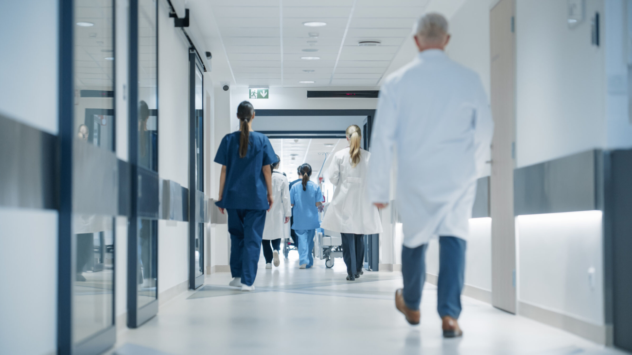 Hospital Hallway with Doctors, Nurses and Specialists in Hospital. Female and Male Physicians, Surgeons, Healthcare Officials Walk Together in Corridor with Their Back to Camera